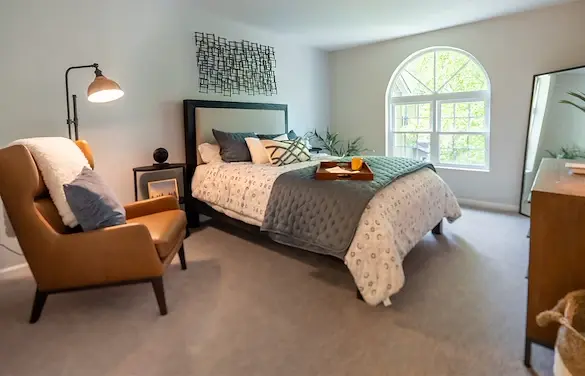 Furnished bedroom at Bellingham at West Chester with a bed, upholstered armchair, bedside table, and arched window.
